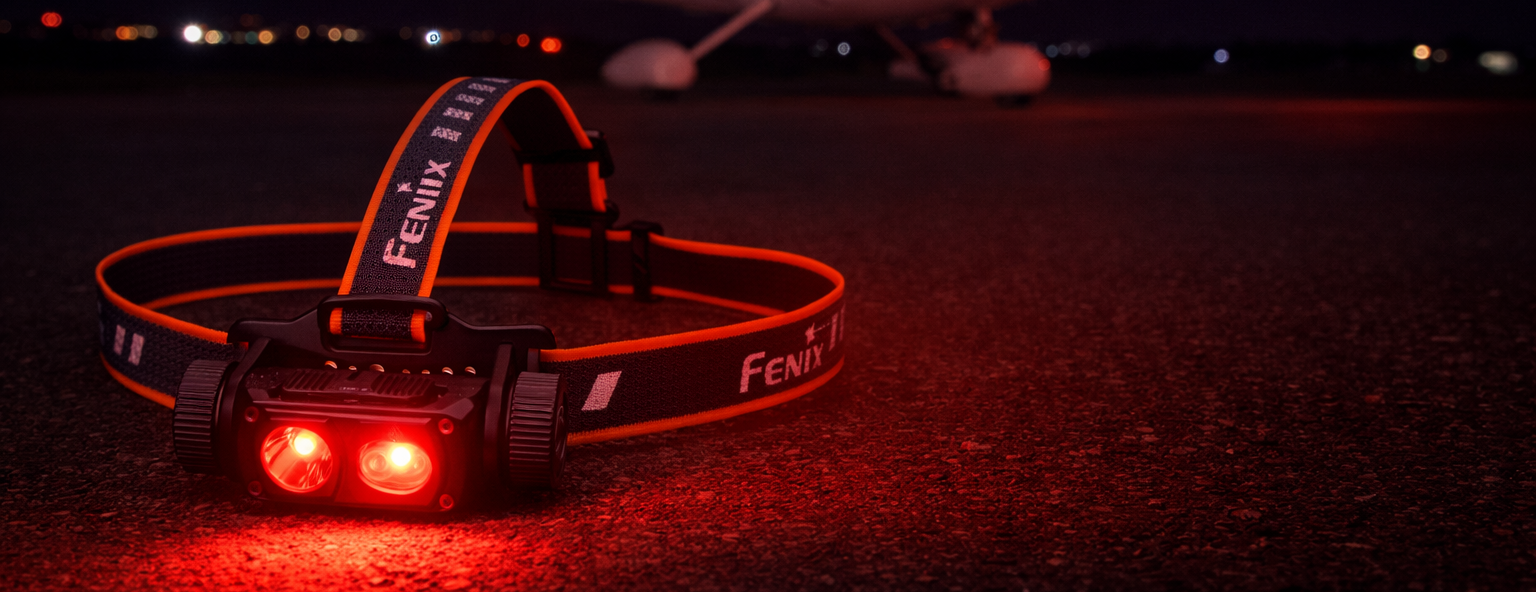 A FENIX headlamp with bright red lights, resting on a dark asphalt surface at night with a blurred propeller airplane in the background.